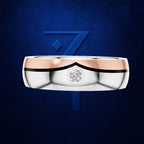 A two-tone wedding band featuring a polished silver body, a rose gold upper band, and a small round-cut diamond set in the center, presented against a textured dark blue background with a stylized blue '7' logo.