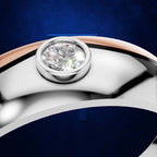 Close-up of a ring with a round brilliant-cut diamond set in a silver-toned band, featuring a rose gold accent along the top curve, against a dark blue textured background.