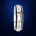 A three-band ring with a white band, a black inner band, and a rose gold outer band, accented with three round-cut diamonds. The ring is shown vertically against a dark blue textured background with a subtle blue geometric logo behind it.