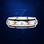 A silver-toned band ring with a polished center, black inlay bands, three clear gemstones, and three rose-gold chevron accents, shown against a dark blue textured background with a blue stylized '7' logo behind it.