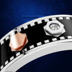 A close-up of a silver-toned ring featuring a black enamel inlay and a filmstrip design, with a rose gold geometric accent and a clear round gemstone in a silver bezel.
