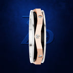 A close-up of a modern ring with a silver-toned outer band, a rose gold center band, and a black inner accent, accented with three round-cut clear gemstones. The ring is shown against a dark textured blue background with a subtle blue geometric logo behind it.