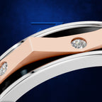 Close-up of a curved ring with a rose gold inlay, a silver outer band, and two round-cut clear gemstones, set against a dark blue textured background.