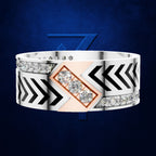 A wide silver-toned ring with black chevron motifs, rows of clear gemstones, and a central rose-gold diamond-shaped inlay featuring three round clear stones, shown on a dark blue textured background.
