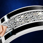 Close-up of a curved ring featuring a row of clear round gemstones between polished silver-toned bands with bold black inlay, set against a deep blue background.