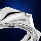 A close-up studio shot of a polished silver-toned ring with a curved row of clear gemstones along the band, set against a deep blue textured background.