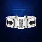 A silver-toned ring with a rectangular clear center stone and small clear stones along the shoulders, set against a dark blue textured background with a blue stylized '7' logo behind it.