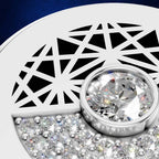 Close-up of a silver-toned jewelry piece featuring a large round clear gemstone in a bezel setting, surrounded by smaller sparkling stones and a geometric cutout pattern on a white curved surface.