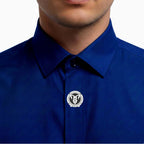 A close-up of a man wearing a royal blue collared shirt, featuring a silver-toned circular lapel pin with a black ram's head on the left chest.