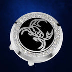 A circular silver-toned accessory with a black center featuring a stylized silver scorpion graphic, surrounded by a border of clear rhinestones, shown on a dark blue textured background.