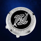 A round silver-toned golf ball marker with a black center featuring a white stylized fish logo, surrounded by a halo of clear rhinestones, set against a dark blue textured background.
