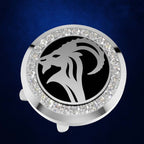 A round silver-toned accessory with a black center featuring a white ram or goat head silhouette, surrounded by a border of clear rhinestones, shown on a dark blue textured background.