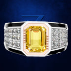 A men's statement ring featuring a large yellow emerald-cut center stone, flanked by rows of clear pavé stones on a wide silver-toned band with black accents, set against a dark blue textured background.