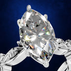 A close-up of a silver-toned ring featuring a large marquise-cut clear gemstone in a prong setting, shown against a dark blue textured background.