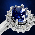 A close-up studio shot of a silver-toned ring featuring a large round brilliant-cut deep blue sapphire center stone surrounded by a halo of small clear round-cut diamonds, set against a dark blue background.