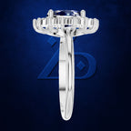 A silver-toned solitaire ring featuring a round brilliant-cut blue gemstone in a prong setting, shown against a textured dark blue background with a subtle logo.