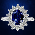An oval-cut blue sapphire ring set in a halo of round-cut clear diamonds on a polished silver band, presented against a dark blue textured background.