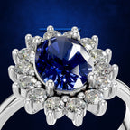 A close-up shot of a silver-toned ring featuring a large oval-cut deep blue gemstone surrounded by a halo of round clear stones, set against a dark blue textured background.