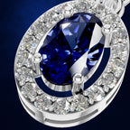 A close-up of a silver-toned oval pendant featuring a large deep blue marquise-cut center stone surrounded by a halo of clear round and round-cut stones, set against a dark blue background.