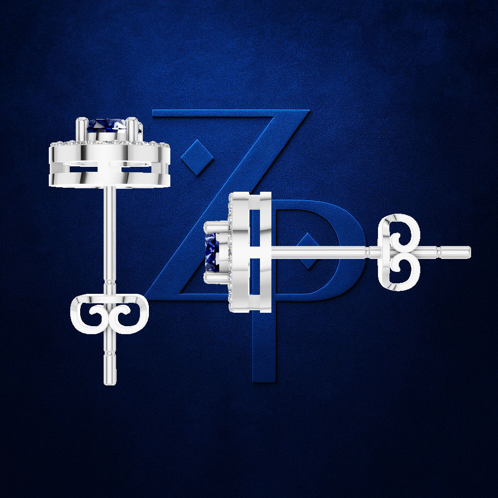 A pair of silver-toned stud earrings with a small dark blue gemstone, shown from front and side angles against a textured dark blue background with a stylized 'ZP' logo.