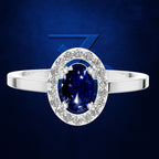 An oval halo ring featuring a deep blue sapphire center stone surrounded by a halo of small clear stones, set on a polished silver band, against a dark blue textured background.
