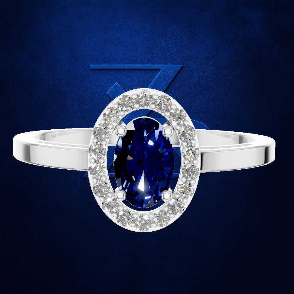 An oval halo ring featuring a deep blue sapphire center stone surrounded by a halo of small clear stones, set on a polished silver band, against a dark blue textured background.