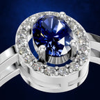A close-up of a silver-toned ring featuring a large oval-cut deep blue gemstone in a prong setting, surrounded by a halo of small clear stones, set against a dark blue background.