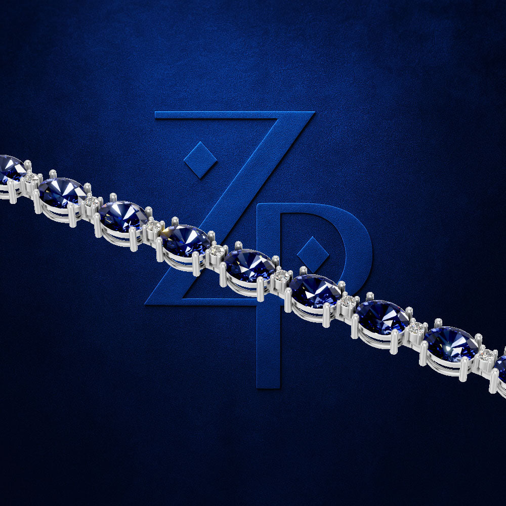 A close-up of a silver-toned tennis bracelet with oval deep blue gemstones, set against a textured dark blue background with a large embossed 'ZP' logo.