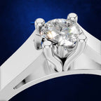 A close-up studio shot of a silver-toned solitaire ring with a large round brilliant-cut clear gemstone, set against a dark blue textured background.