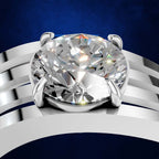 A close-up studio shot of a solitaire engagement ring featuring a large round brilliant-cut diamond set in a silver-colored metal band with multiple curved parallel prongs, against a dark blue textured background.
