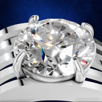 A close-up studio shot of a silver-toned ring with a large oval clear gemstone in a prong setting, shown against a dark blue background with a blurred metallic band.