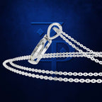 A silver-toned cremation urn pendant with a faceted crystal-like front, shown on a matching chain against a dark blue textured background with a large stylized blue monogram behind it.