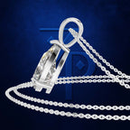 A silver-toned pendant necklace with a clear faceted gemstone in a polished bezel setting, shown with a matching chain on a dark blue textured background with a large blue monogram behind it.