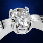 A close-up studio shot of a silver-toned ring with a large oval-cut clear gemstone in a prong setting, shown against a deep blue background.