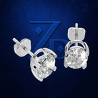 A pair of silver stud earrings with round clear gemstones in a prong setting, shown from two angles against a dark blue textured background with a faint stylized 'ZPD' logo.