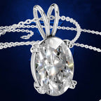 A silver-toned oval pendant necklace with a large clear faceted gemstone, shown on a dark blue background. The pendant is suspended from a delicate chain that loops around the top of the bail.