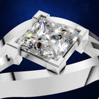 A close-up studio shot of a silver-toned ring with a large clear princess-cut diamond. The diamond is set in a square four-prong setting on a polished band, shown against a dark blue background.