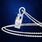 A silver-toned pendant necklace with a geometric, shield-like clasp and a clear faceted stone, shown on a dark blue textured background with a large stylized blue letter behind it.
