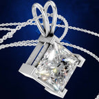 A silver-toned diamond pendant necklace featuring a princess-cut clear diamond in a four-prong setting with a heart-shaped bail, shown on a dark blue textured background.
