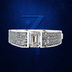 A silver-toned ring with a large rectangular clear center stone and textured gray inlays on both sides, shown against a dark blue background with a large stylized blue number 7 behind it.