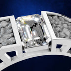 A close-up of a white metal ring setting featuring a large, brilliant-cut clear center stone, with textured gray side stones set in a geometric band against a dark blue background.