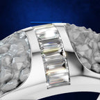 A close-up of a silver-toned ring featuring a central channel set with multiple baguette-cut clear stones, flanked by textured, hammered silver sides, set against a dark blue background.
